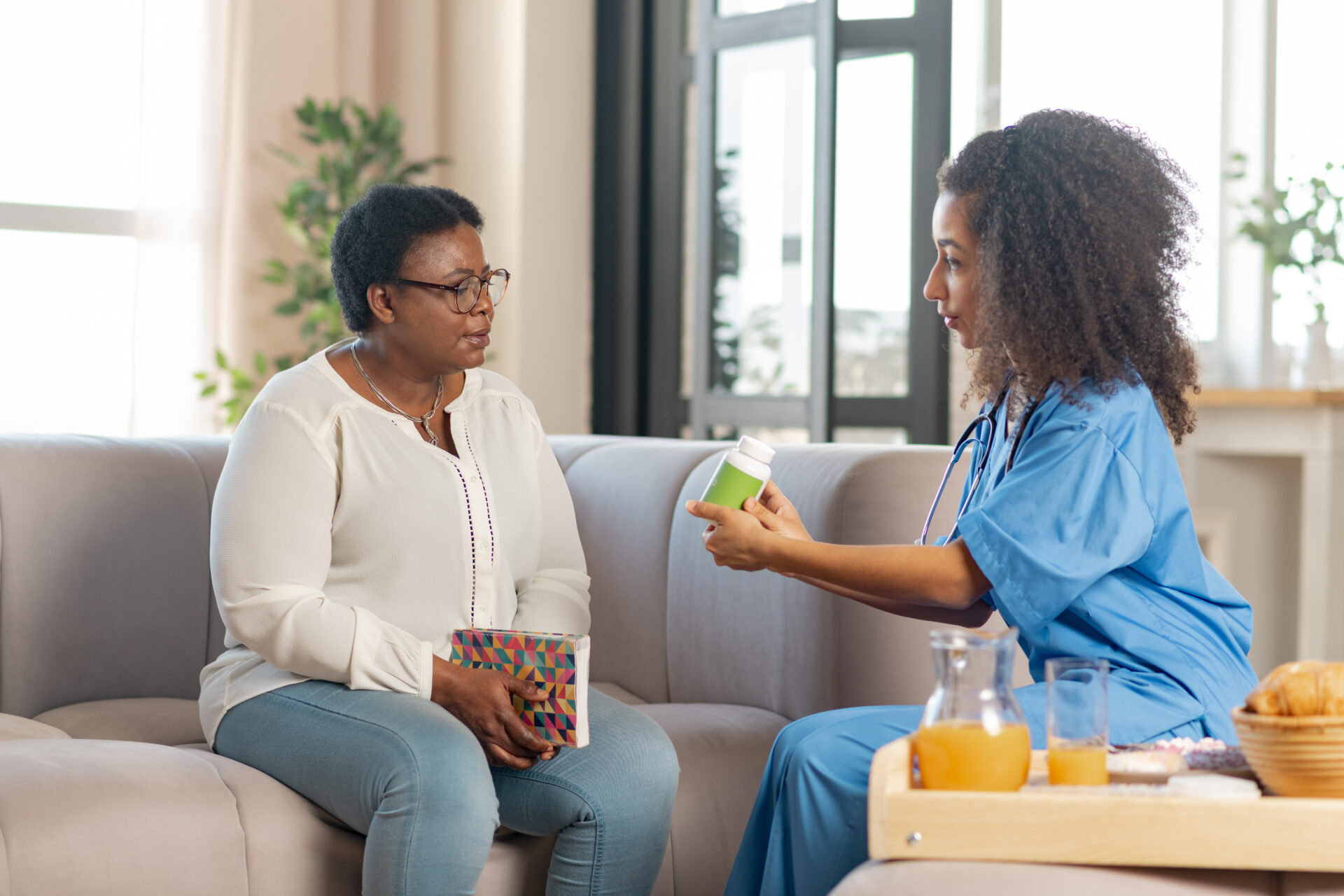 Giving some pills. Sick dark-skinned elderly lady looking at caring young nurse giving her some pills